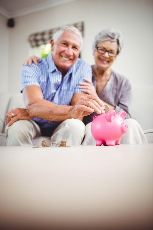 Portrait of senior man sitting with woman on sofa and putting coins in piggy bank