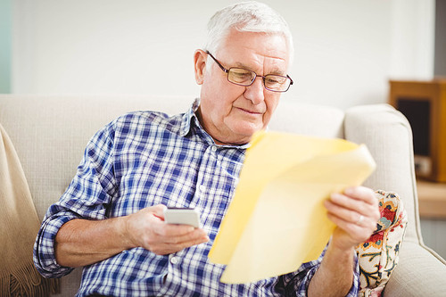 Senior man looking at a document and using mobile phone in living room
