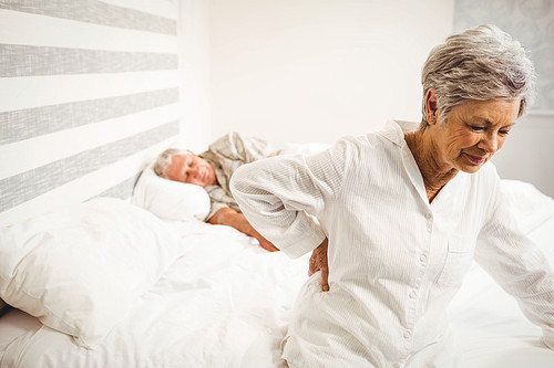 Senior woman suffering from backache sitting on bed in bedroom
