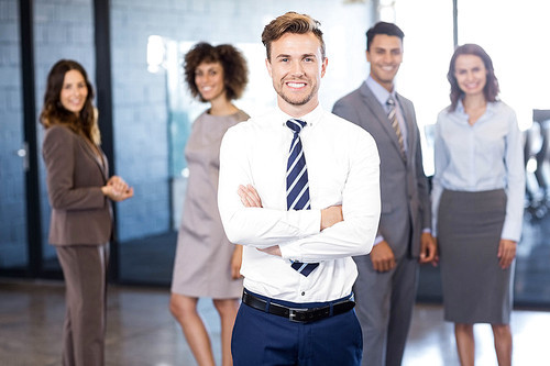 Successful businessman smiling at camera with his arms crossed and his colleagues posing in background