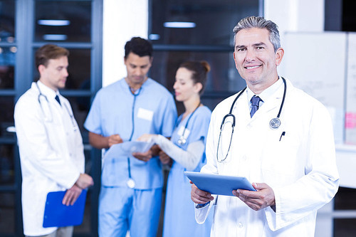 Doctor holding digital tablet and smiling at camera while his colleagues discussing in background