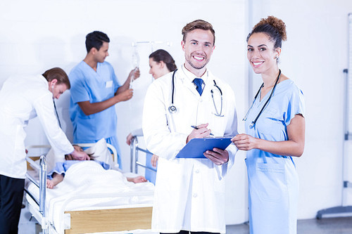 Portrait of doctors holding clipboard and smiling at camera while other doctor examining a patient behind in hospital