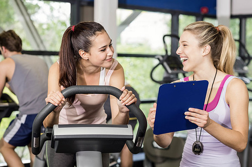 Trainer woman talking with a woman doing exercise bike at gym