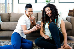 Smiling lesbian couple sitting on rug and looking at their mobile phone in living room