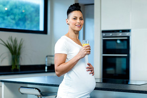 Portrait of pregnant woman standing with hand on stomach and drinking juice in kitchen