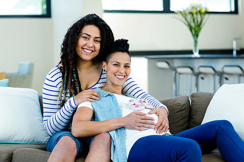 Pregnant lesbian couple sitting on sofa with a pair of pink baby shoes