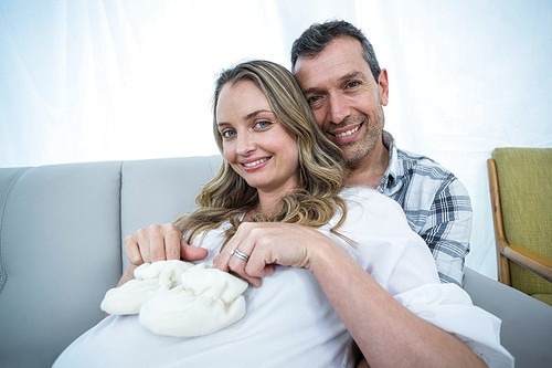 Pregnant couple sitting on sofa with baby shoes on belly in living room