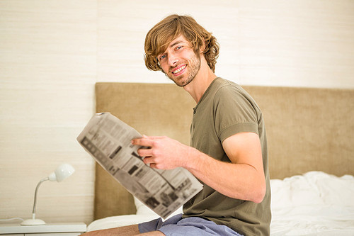 Handsome man reading a newspaper sitting on the edge of his bed