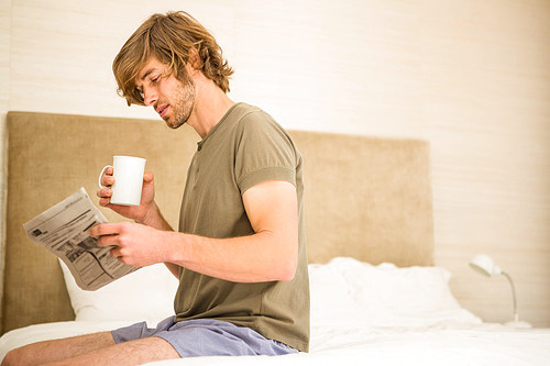 Handsome man reading the news and drinking coffee sitting on his bed