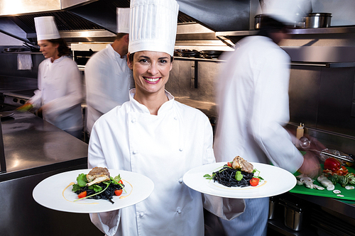 Happy chef presenting her food plates in the commercial kitchen while team working behind her