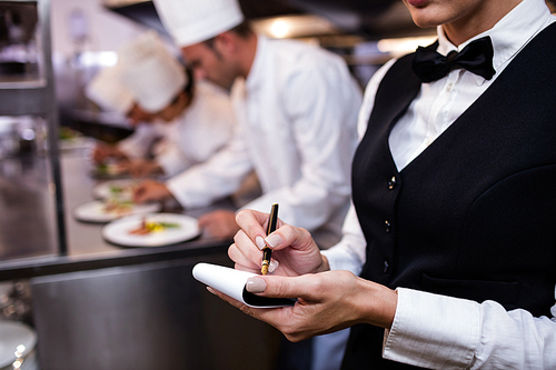 Close-up of waitress with notepad in commercial kitchen and chefs preparing food in background