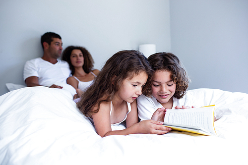 Brother and sister reading book together on bed while parents relaxing in background
