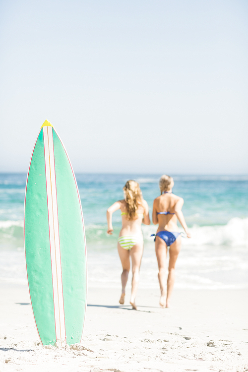 Surfboard in sand and two rear women running on the beach on a sunny day