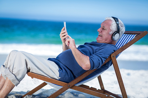 Mature man resting on a deck chair listening to music with smartphone on the beach