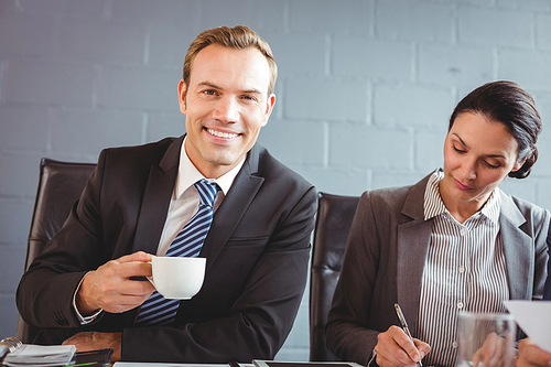 Businessman holding cup and smiling at camera and his colleague working beside him