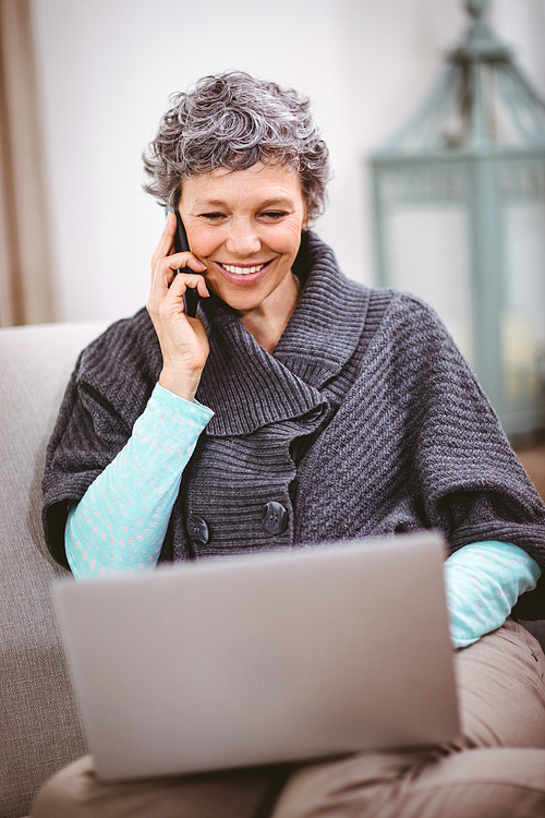 Smiling mature woman with laptop using mobile phone while sitting on sofa at home