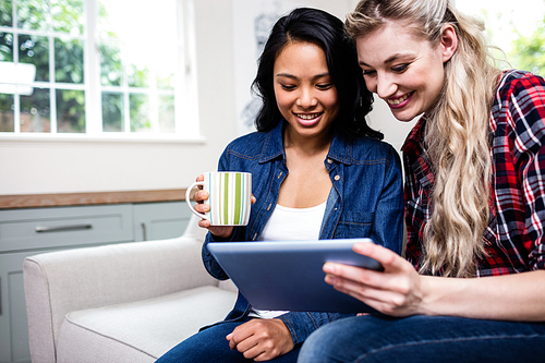 Close-up of young female friends with cup and digital tablet sitting on sofa at home