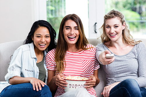 Portrait of cheerful female friends with remote and popcorn sitting on sofa at home