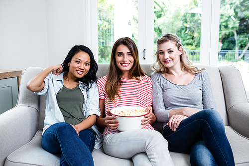 Portrait of cheerful young female friends with popcorn sitting on sofa at home