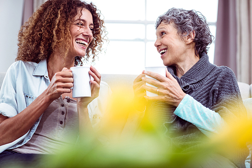 Smiling mother and daughter holding coffee mugs while discussing at home