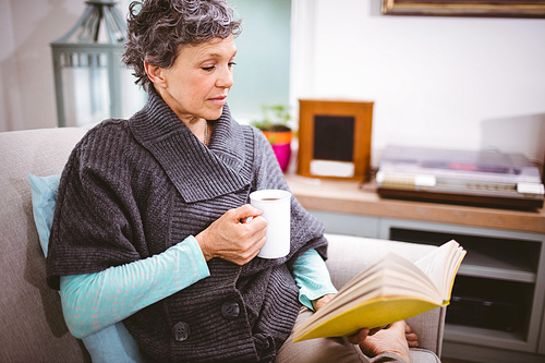 Mature woman reading book and holding coffee mug while sitting at home