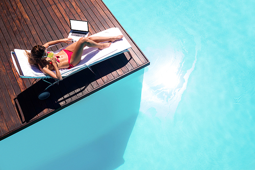 Woman drinking a cocktail and using laptop by swimming pool