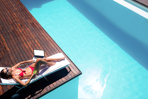 Woman drinking a cocktail and using laptop by swimming pool