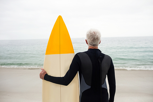 Rear view of senior man in wetsuit holding a surfboard on the beach