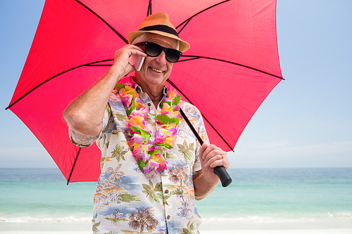 Senior man holding umbrella while talking on phone at beach