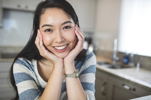 Portrait of happy woman sitting in kitchen with hand on chin