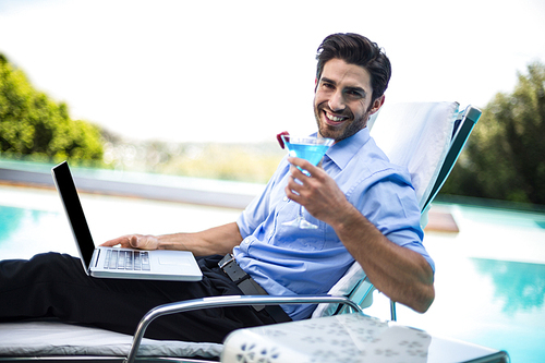 Smart man having martini and using laptop while relaxing near pool