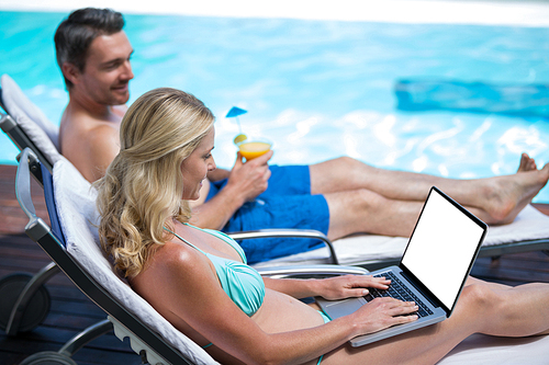 Couple sitting near pool with a laptop and martini glass