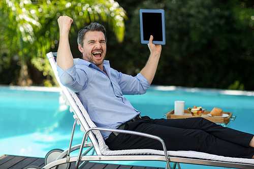 Excited man relaxing on sun lounger and holding a digital tablet near the pool