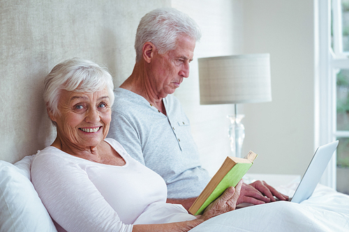 Portrait of smiling senior woman reading book while man using laptop on bed