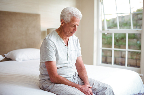 Worried senior man looking down while sitting on bed in room