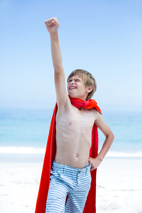 Shirtless boy in superhero costume with hand raised standing at beach