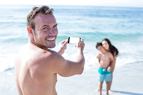 Portrait of man smiling while photographing wife and son at sea shore