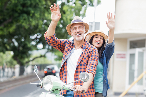 Portrait of happy couple waving hand while sitting on motor scooter