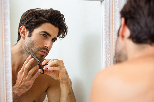 Smart young man cutting beard with scissor in front of bathroom mirror