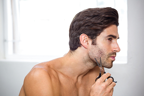 Young man cutting beard with scissor in bathroom