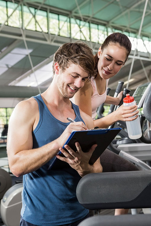 Trainer writing on a clipboard while woman exercising on a elliptical machine at gym