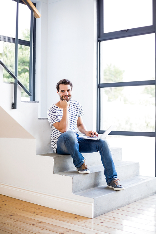 Portrait of happy man sitting on steps using laptop at home