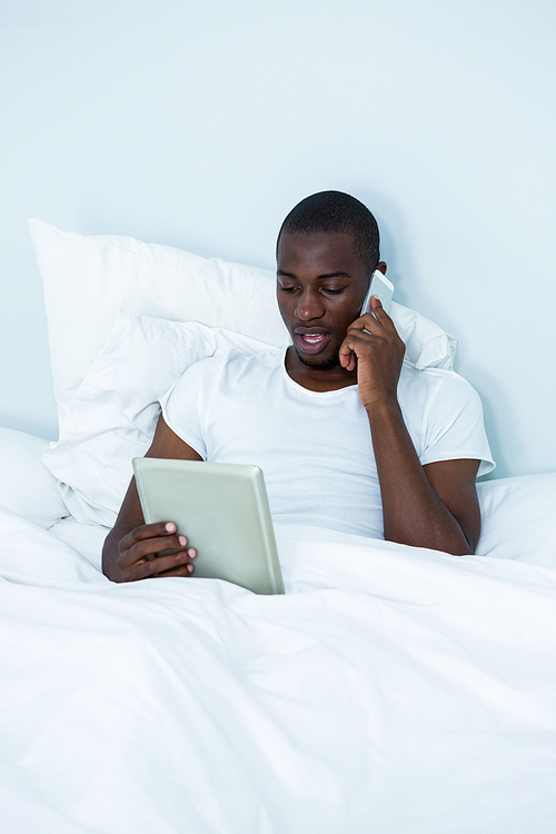 Man looking at digital tablet while talking on phone in bedroom