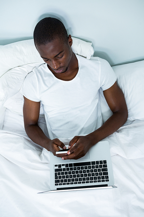 Young man using laptop and phone on bed in bedroom