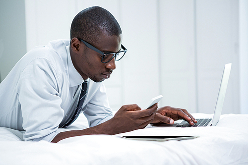 Young man using laptop and phone on bed in bedroom