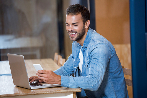 Man text messaging on phone while sitting at desk with laptop in office