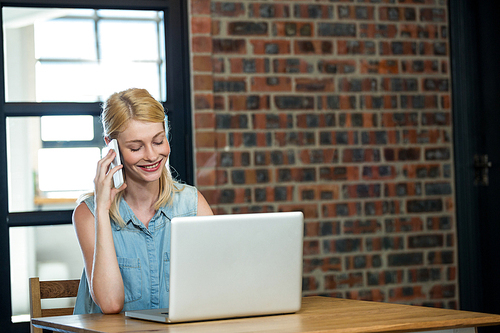 Woman talking on phone while using laptop in office