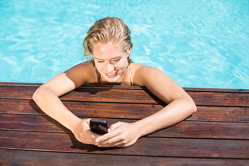 Beautiful woman leaning on poolside and typing a text message on cellphone