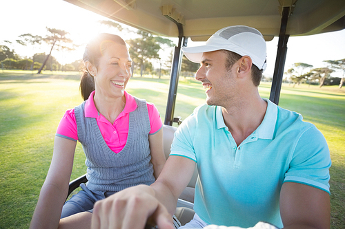 Smiling golfer couple looking at each other while sitting in golf buggy on sunny day