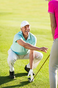 Smiling mature golfer man crouching while teaching woman standing on field